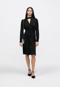 Rochie neagră cu mâneci lungi, decolteu adânc în V și cordon în talie. Textură netedă, mătăsoasă; asortată cu pantofi negri cu toc și o geantă argintie.