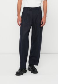 Dark blue pinstriped trousers, wide-leg fit, textured fabric, secured with a button, paired with black shoes. Simple, tailored design.