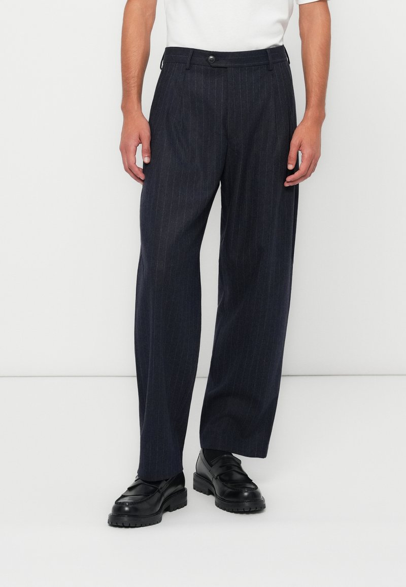 Dark blue pinstriped trousers, wide-leg fit, textured fabric, secured with a button, paired with black shoes. Simple, tailored design.
