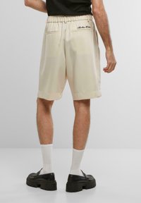 Shorts beiges avec une texture lisse, une taille élastique et des poches arrière. Ourlet retourné et détail de broderie noire sur la taille.