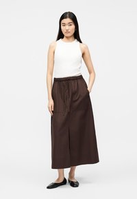 Jeune femme aux longs cheveux raides, portant un haut blanc sans manches, une jupe midi marron avec cordon de serrage, et des ballerines noires, debout la main dans la poche.