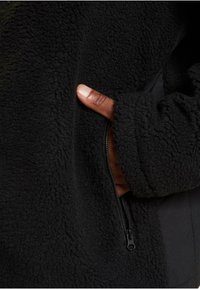 Hand in der Tasche einer schwarzen Fleecejacke mit sichtbarem schwarzen Reißverschluss, zeigt strukturierter Stoff und teilweise herausragenden Daumen aus dem Ärmel.