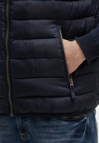 Gilet imbottito blu navy con sezioni trapuntate, tasca laterale con zip e zip color argento. Indossato sopra una maglietta a maniche lunghe blu navy e jeans blu.