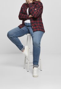 Röd och svart rutiga skjorta över en vit topp, tillsammans med blå jeans och vita sneakers. Sitter på en pall med en avslappnad pose.