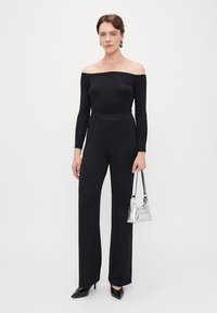 Zwarte off-the-shoulder top gecombineerd met gestructureerde, hoge zwarte broeken. Model houdt een kleine zilveren handtas vast en draagt zwarte hakken.