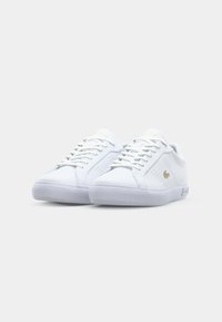 Baskets en cuir blanc à texture lisse, design à lacets, accent logo doré, et semelle en caoutchouc translucide. Style athlétique classique.