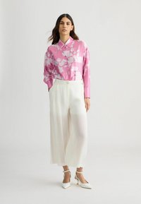Roze bloempatronde blouse met een kraag, gecombineerd met hooggetailleerde witte wijde broek en witte puntige hakken, tegen een neutrale achtergrond.