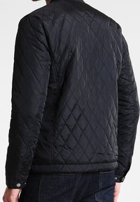 Quiltet sort nylonjakke med diamantmønster, tekstureret overflade, elastiske manchetter og trykknapper i kanten, med et enkelt kravedesign.