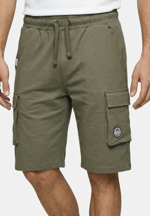 Homme portant un short cargo vert olive avec taille à cordon et poches à rabat, debout devant un fond uni.