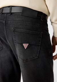 Jean en denim noir avec une finition texturée. Comprend un patch logo triangulaire rouge et blanc sur la poche arrière et une ceinture en cuir noir avec branding.