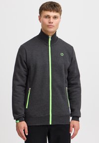 Chaqueta de felpa gris oscuro con cremallera y cuello alto, que presenta detalles en verde en la cremallera y el logo, dos bolsillos laterales y puños de canalé.