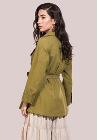 Chaqueta verde oliva con cintura ceñida, mangas largas y tejido ligero. Presenta una abertura en la parte posterior y un corte relajado. Superpuesta sobre una falda crema.