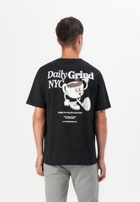 Svart bomull t-skjorte med en tegneserie-kop kaffe grafikk på baksiden, med hvit tekst "Daily Grind NYC" og nettstedsdetaljer.