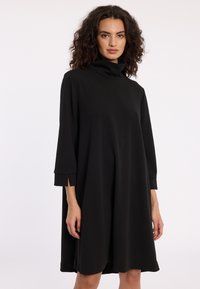 Vrouw met krullend haar, gekleed in een losse zwarte coltrui jurk met driekwart mouwen en zijsplitten, staande tegen een effen achtergrond.