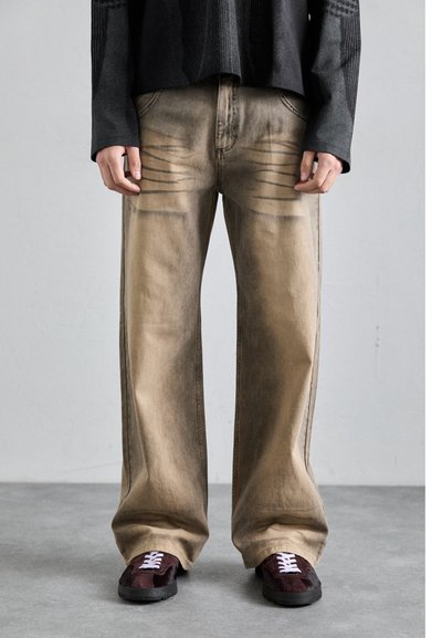 Beige weit geschnittene Jeans mit verblasster Textur und horizontalen Linien auf der Vorderseite. Kombiniert mit dunklen Sneakers mit weißen Schnürsenkeln.