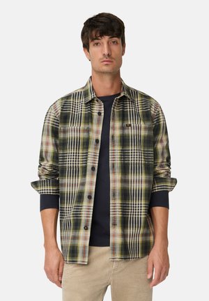 Man in een groen, zwart en wit geruit overhemd over een zwart longsleeve shirt met beige broek, staand tegen een effen witte achtergrond.