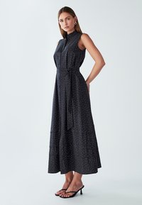 Czarna bezrękawnikowa sukienka z kołnierzykiem, o warstwowym kroju, w białe kropki, z paskiem w talii i krojem A-linii; zestawiona z czarnymi sandałkami na obcasie.