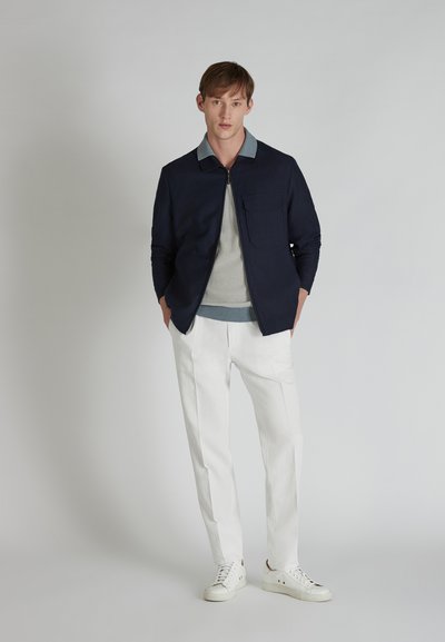 Chaqueta de cremallera azul marino con un cuello azul claro, suéter de punto gris debajo, pantalones blancos y zapatillas blancas. Tejido suave con líneas limpias.