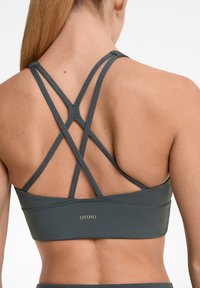 Tamno siva sportska grudnjak s dizajnom križnog leđa. Izrađena od glatkog, rastezljivog materijala s detaljem logotipa na leđima.