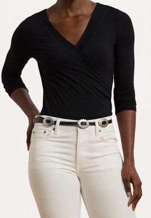 Femme portant un haut noir style portefeuille avec manches trois-quarts et un jean blanc taille haute avec une ceinture noire avec trois boucles argentées.