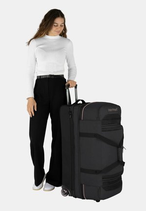 Schwarze Reisetasche auf Rädern mit pinken Akzenten. Verfügt über mehrere Taschen, strapazierfähigen Stoff und einen ausziehbaren Griff. Steht aufrecht neben einer Person.