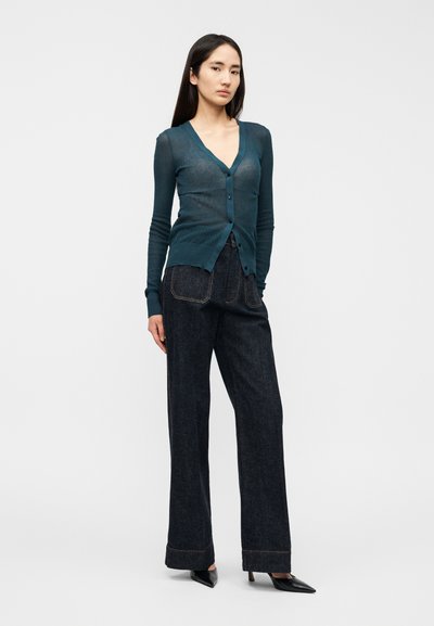 Πλεκτή μπλετυρκουάζ ζακέτα με βαθύ V λαιμόκοψη και κλείσιμο με κουμπιά, σε συνδυασμό με ψηλόμεσο σκουρόχρωμο τζιν παντελόνι. Φόρμα που αγκαλιάζει τη σιλουέτα με μακριά μανίκια.