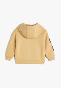 Felpa con cappuccio di colore beige chiaro, dal texture morbido, vestibilità oversize, polsini e fondo a coste, e dettaglio testuale stampato in nero sulla manica.