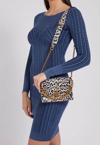 Kabelka s leopardím potiskem a texturovaným povrchem, s přívěskem zlatého řetízku a logem, držená proti modrým žebrovaným šatům.