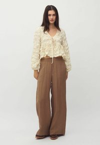 Krémový, textúrovaný crop top s viazaním vpredu, skombinovaný s voľnými, hnedými nohavicami s vysokým pásom. Oblečenie má uvoľnené, prúdiace línie.