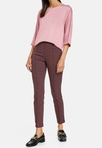 Blusa di raso rosa con maniche larghe, abbinata a pantaloni aderenti con motivo geometrico in rosso, nero e bianco. Mocassini neri con accenti dorati.