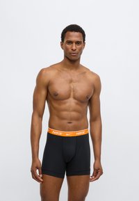 Fekete boxeralsó narancssárga Nike logóval a derékpánton. Sima anyag és testhez simuló kialakítás, középhosszúságú, a comb közepéig érő szabással.