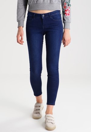 Kvinde iført mørkeblå skinny jeans, lys grå sweatshirt med blomsterbroderi på ærmet og beige velcro sneakers.