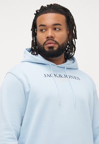 Man with dreadlocks and beard wearing a light blue hoodie with "JACK & JONES" text, looking to his left against a plain background.