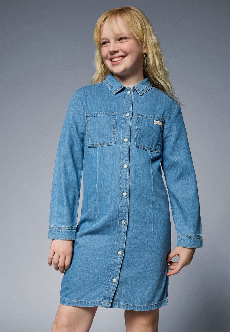 Ljusblå denimklänning med knäppning framtill, långa ärmar och två bröstfickor. Har en krage och rak silhuett.