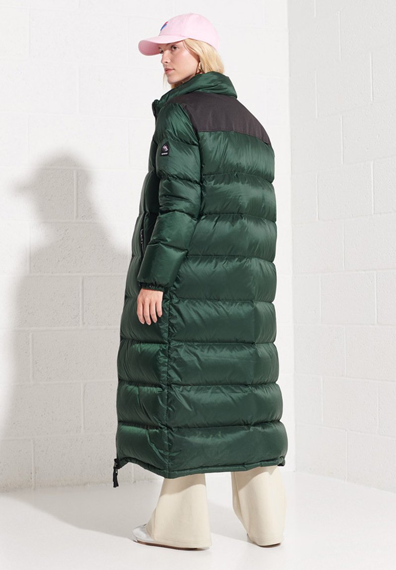 Długa zielona kurtka puchowa o błyszczącej fakturze, z czarną górną częścią, bocznymi zamkami i wysokim kołnierzem. Noszona z różową czapką.