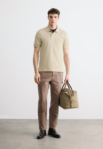 Jeune homme debout portant un polo beige, un pantalon marron et des chaussures foncées, tenant un sac de voyage kaki dans sa main droite.