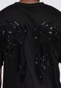 Musta t-paita, jossa on suuri, helmin koristeltu siipikuvio selässä. Siivet on koristeltu mustilla paljeteilla ja yksityiskohtainen ompelu luo rakennetta ja vivahteita.