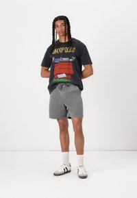 T-shirt graphique noir avec un design coloré de Snoop Dogg, associé à un short gris. Le modèle porte des chaussettes blanches et des chaussures de sport noires avec des rayures.