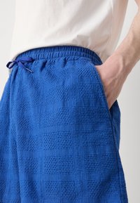 Pantalones cortos azules con textura y patrón de cuadrícula, cinturilla elástica y bolsillos laterales. Fabricados con un material suave. Detalles con cordón en la cintura.