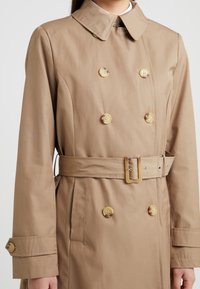 Kvinna som bär en beige dubbelknäppt trenchcoat med matchande bälte, knäppta ärmslut och en klassisk krage mot en enfärgad bakgrund.