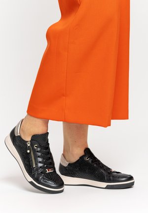 Füße in schwarzen, strukturierten Ledersneakern mit Reißverschlüssen neben orangefarbenen, weit geschnittenen Hosen auf weißem Hintergrund.