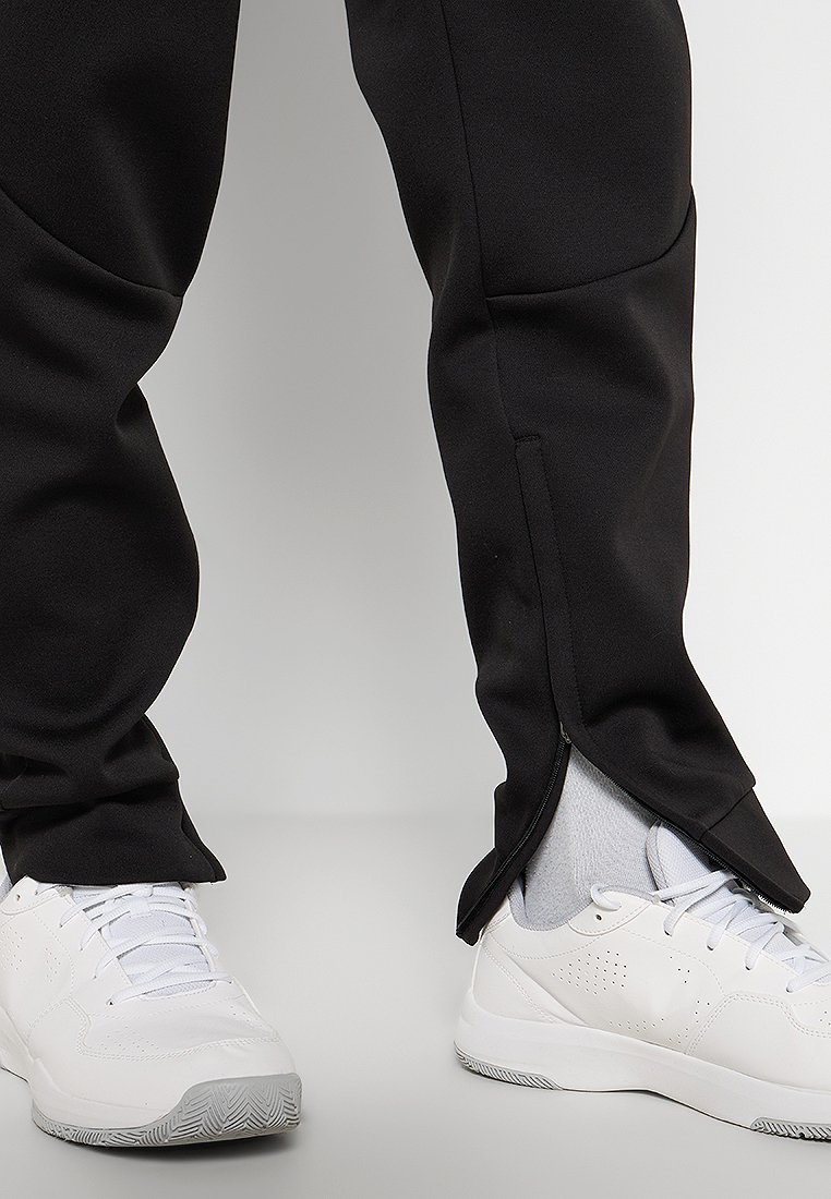 Pantalons de sport noirs avec un design fuselé, dotés d'ourlets zippés. Associés à des chaussures de sport blanches pour un look contrasté.