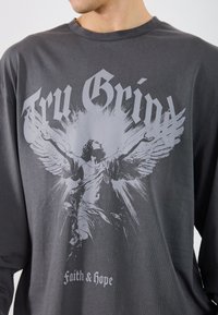 Siva majica dugih rukava sa velikim grafičkim printom figure s krilima, uz tekst "Try Grind" i "Faith & Hope" u kontrastnim bojama.