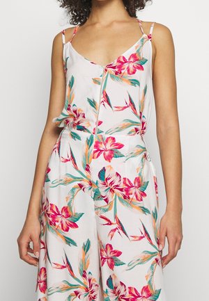 Woman wearing a white sleeveless jumpsuit with bright pink, orange, and green tropical floral patterns and thin shoulder straps.