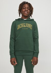 Sudadera verde con texto dorado "JACK&JONES" y "intl. desde 1990". Cuenta con un bolsillo frontal y una tela texturizada. Combinada con pantalones joggers a juego.