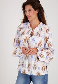 Langemouwige blouse van witte stof met een blauw en bruin abstract patroon, met een gerimpelde halslijn en gathered manchetten. Draag met jeans.