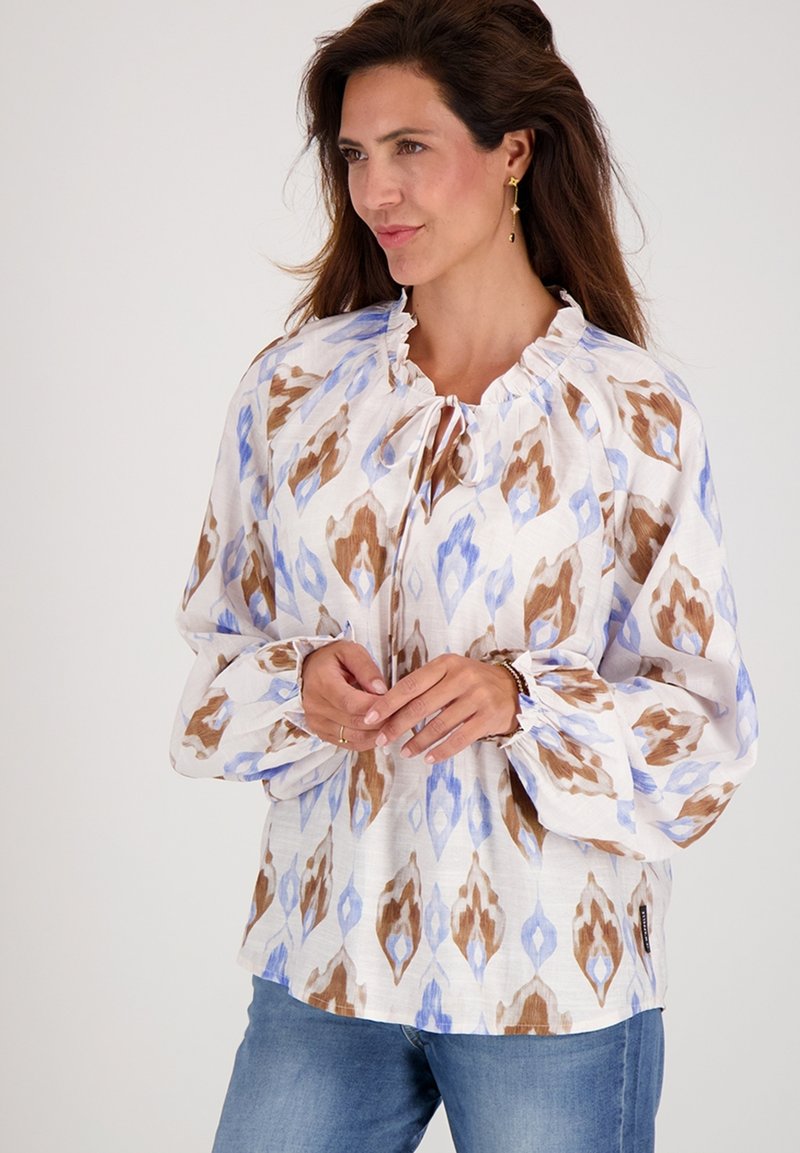 Langemouwige blouse van witte stof met een blauw en bruin abstract patroon, met een gerimpelde halslijn en gathered manchetten. Draag met jeans.