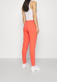 Kvinna i korallfärgade joggingbyxor, vit tanktop och vita sneakers, stående mot en ljusgrå bakgrund, bakifrån.