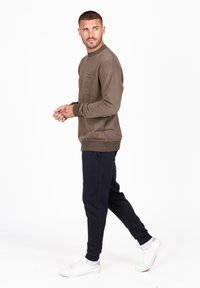 Brązowy sweatshirt z kieszenią na przodzie i ciemnymi mankietami, w połączeniu z granatowymi joggerami i białymi sneakersami, na tle białego tła.
