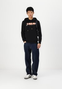 Sudadera con capucha negra y logo en contraste, jeans de mezclilla y zapatillas blancas con acentos negros. El modelo está de pie contra un fondo liso.